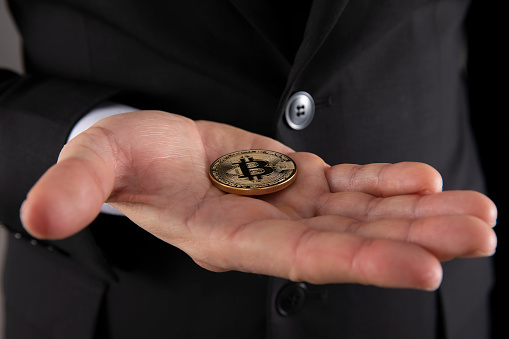 Istanbul, Turkey - February 20, 2022: Hand of Businessman in a suit is holding a gold colored memorial coin of bitcoin in his palm