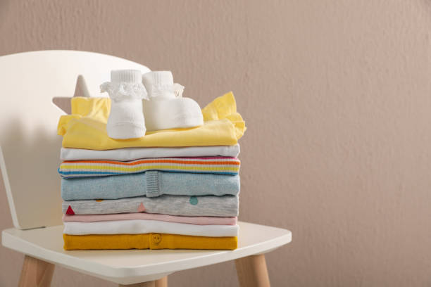 Stack of baby clothes and booties on chair near beige wall. Space for text Stack of baby clothes and booties on chair near beige wall. Space for text baby clothing stock pictures, royalty-free photos & images