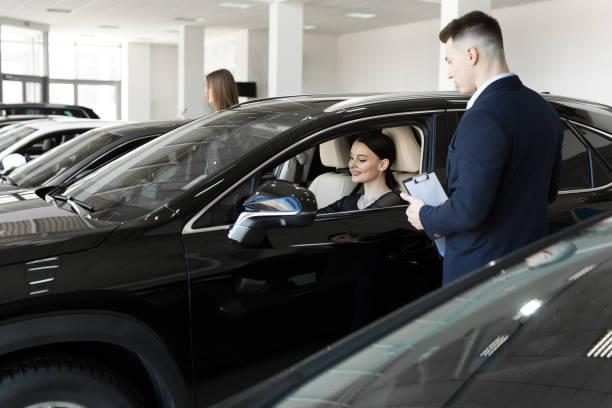 Side view of young beautiful woman sitting inside car and holding hand on steering wheel. She smiling and talking with manager of car dealership. Car agent representing inside of automobile. vista lateral de una joven hermosa mujer sentada dentro del automóvil y tomándose de la mano en el volante. ella sonriendo y hablando con el gerente del concesionario de automóviles. agente de automóviles que representa el interior del automóvil