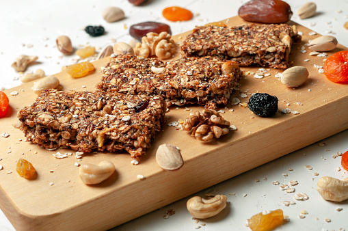 bar with granola, dried fruits and nuts on a light wooden board with dried fruits, nuts and cereals scattered nearby, close-up.