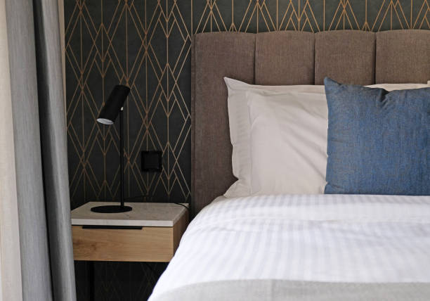 istock Close-up fragment of bedroom with empty bedside table, reading lamp and a socket in modern interior​ design home or hotel. Soft pillow and blanket, stylish comfortable furniture 1392114555