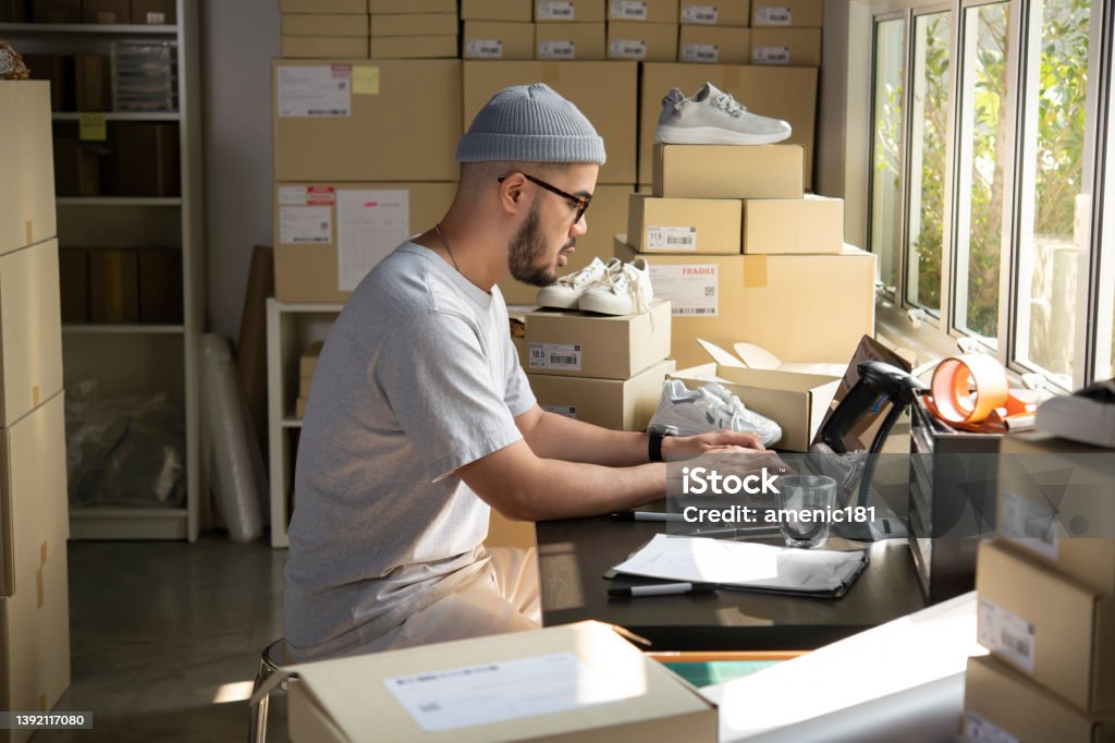 E-commerce male business owner working on laptop computer in store warehouse. Asian man online seller confirming orders from customer on laptop. E-commerce male business owner working on laptop computer in store warehouse. Retail Stock Photo
