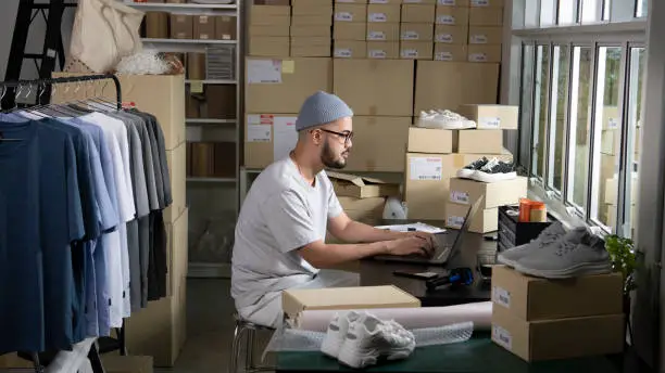 Photo of E-commerce male business owner working on laptop computer in store warehouse.