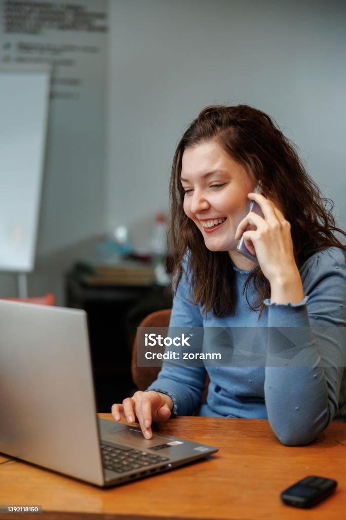 Beautiful post-grad using laptop and phone at the desk in the office Beautiful university student using laptop and talking on the smart phone at the desk in the office 30-34 Years Stock Photo