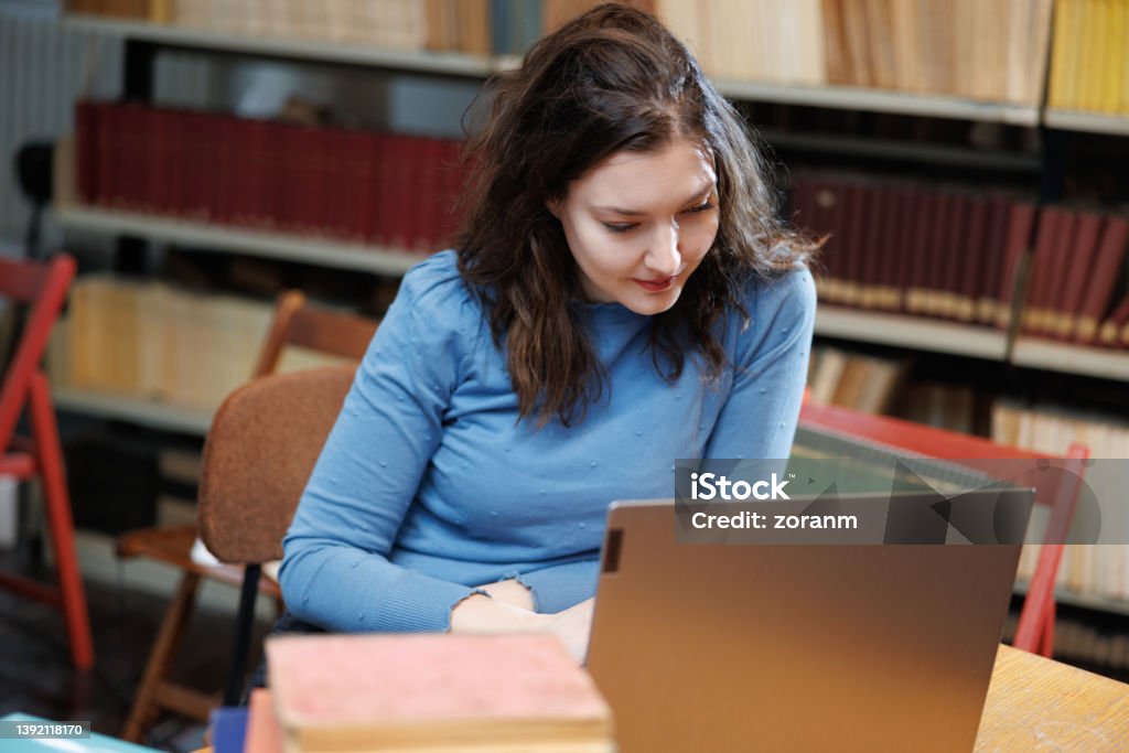 Female post-grad using laptop at the desk in university library Beautiful university student using laptop at the desk with a stack of selected books in university library 30-34 Years Stock Photo