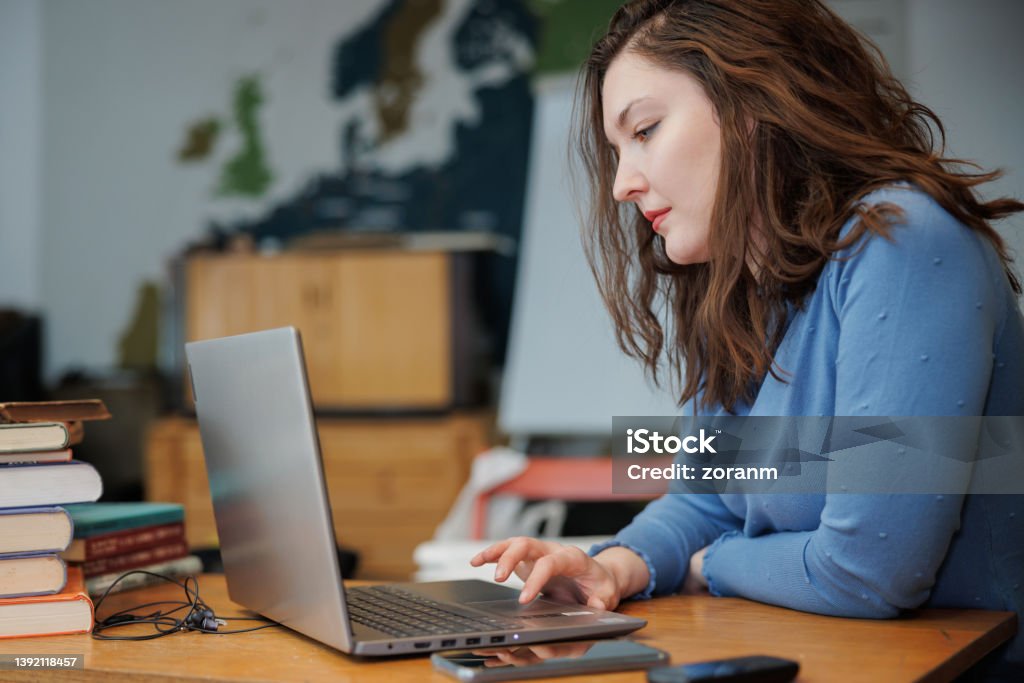 Female post-grad using laptop at the desk with stacked books Beautiful university student sitting in the office at the desk with stacked books and using laptop Library Stock Photo