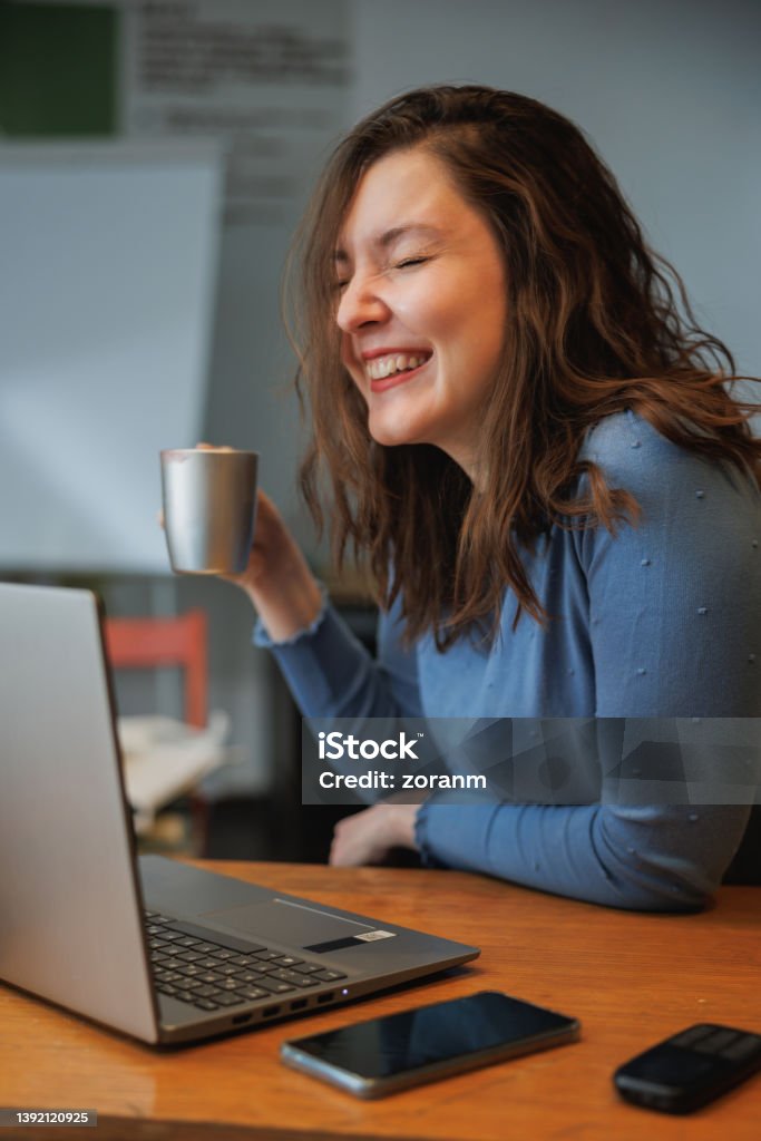 Beautiful post-grad sitting at the desk in the office and having coffee with a smile Female university student sitting at the desk in the office with laptop and smart phone, having coffee on a break and smiling with eyes closed 30-34 Years Stock Photo