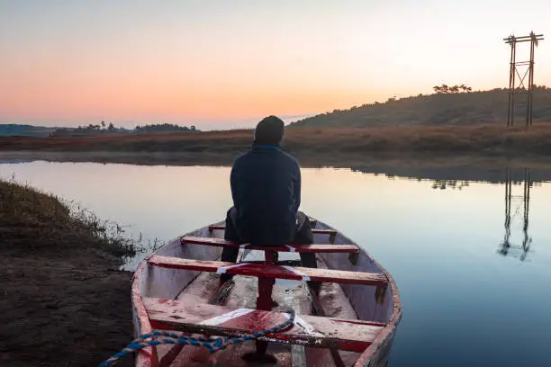 Photo of man sitting at traditional wood boat at calm lake with dramatic colorful sky reflection at morning