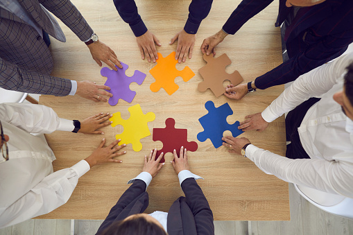 Business team standing around an office table trying to match and fit colorful pieces of a jigsaw puzzle as a metaphor for teamwork, creativity and looking for working business solutions, high angle