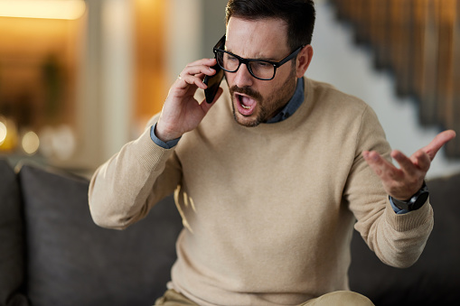 Displeased man arguing with someone over smart phone while working at home.