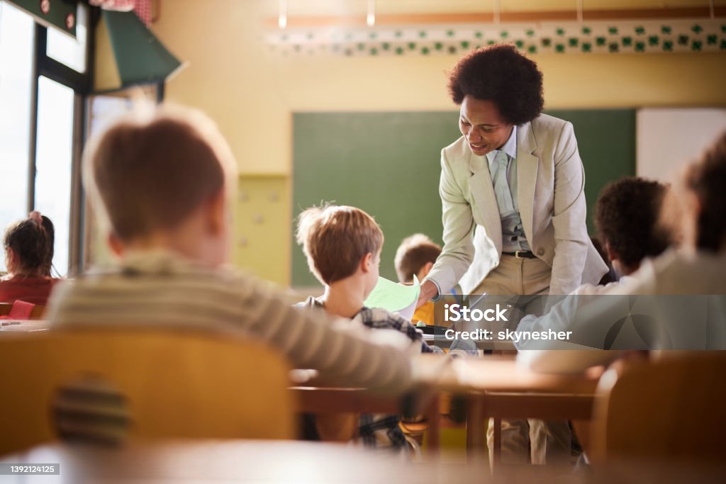 Happy African American teacher assisting her students on a class at elementary school. Happy black professor assisting schoolboy on a class at school. Teacher Stock Photo
