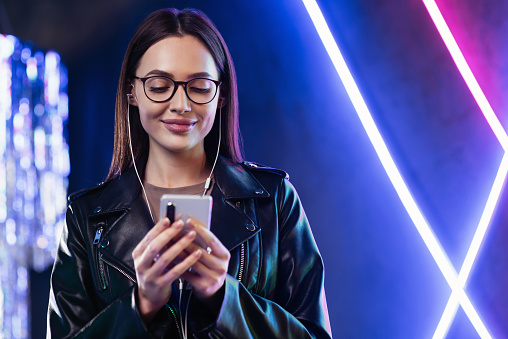 Smiling student girl looking at mobile phone at night club. People, technology, connection, mobile apps, communication and active lifestyle concept