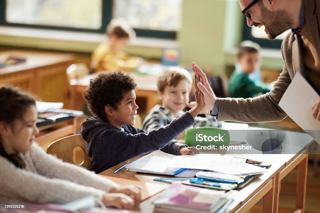 Happy teacher and schoolboy giving each other high-five on a class. Happy male teacher giving high-five to his black elementary student on a class in the classroom. Teacher Stock Photo