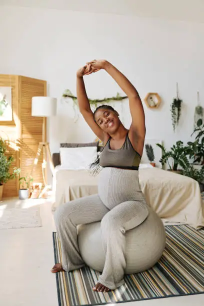 African young woman doing exercises on fitness ball in her bedroom during pregnancy