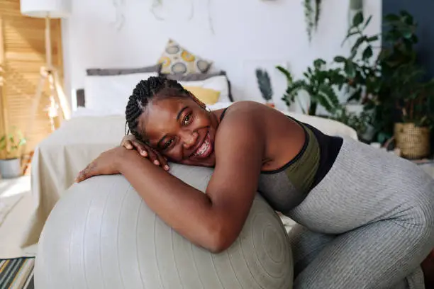 Portrait of happy African woman smiling at camera while exercising with fitness ball during pregnancy
