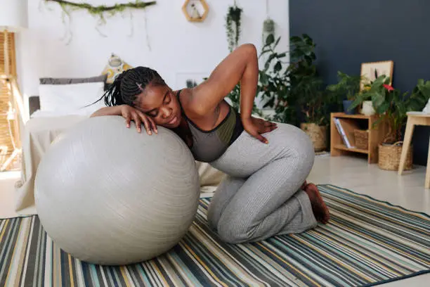African pregnant girl in pain leaning on fitness ball with her eyes closed during labor in room