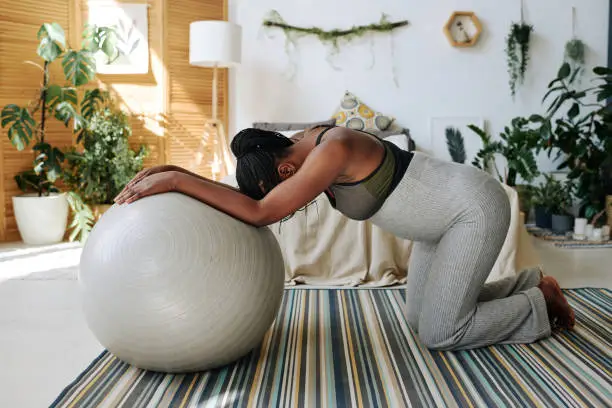 African pregnant woman standing on knees leaning on fitness ball feeling pain during contractions, she giving birth at home