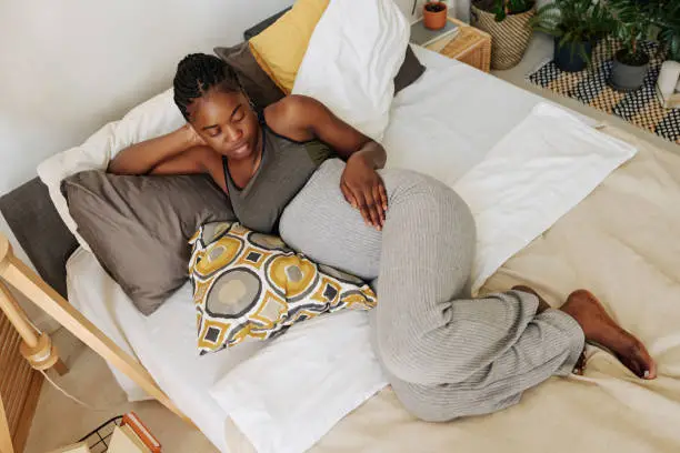 High angle view of African young girl looking at her pregnant belly while relaxing on cushions in bed