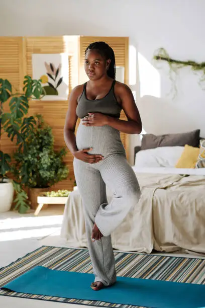 African young woman touching her pregnant belly while standing in yoga pose on exercise mat in her bedroom