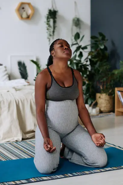African pregnant woman sitting on exercise mat and deeply breathing during meditation at home