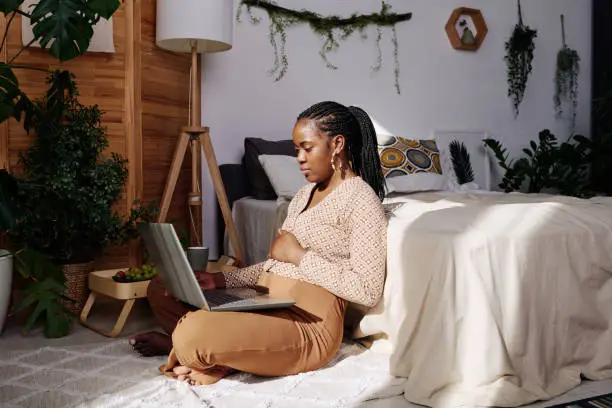 African pregnant woman sitting on floor in bedroom with laptop on her legs having online consultation with doctor
