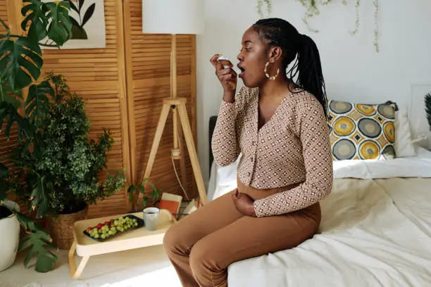 African pregnant girl having an asthma, she sitting on bed in bedroom and using her inhaler