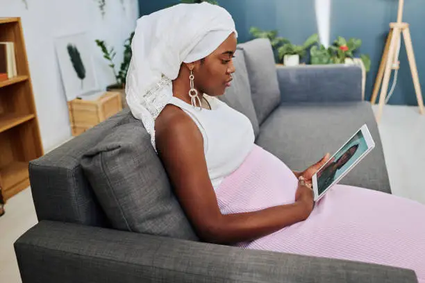 African pregnant girl using digital tablet to talk to her doctor online while sitting on sofa in living room