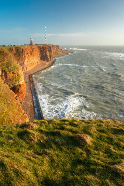 Tall red dramatic cliffs of Heligoland island with rough sea and a lighthouse far in the back. Sunny windy winter day in Helgoland in the North sea, Germany. Tall red dramatic cliffs of Heligoland island with rough sea and a lighthouse far in the back. Sunny windy winter day in Helgoland in the North sea, Germany helgoland stock pictures, royalty-free photos & images