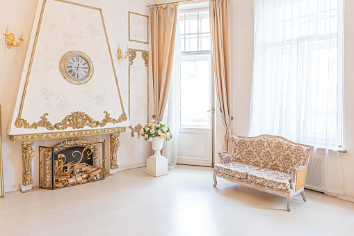 luxurious expensive interior of a large baroque royal living room. antique furniture, gold trim, huge windows, fireplace with gold stucco on the walls. full of daylight