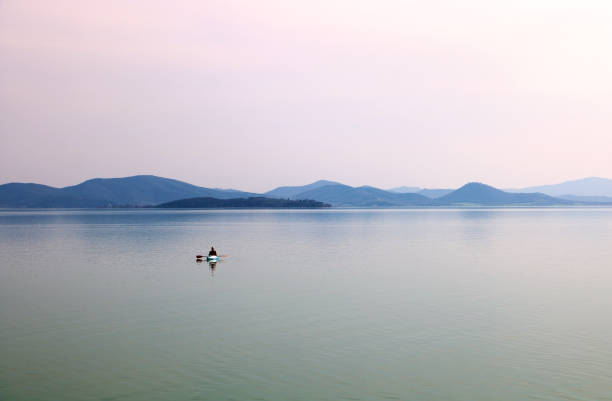The photo was taken at Lake Trasimeno on a beautiful spring day and shows a lonely canoe in the middle of the lake and, behind, one of the islands, and hills and mountains at the horizon.