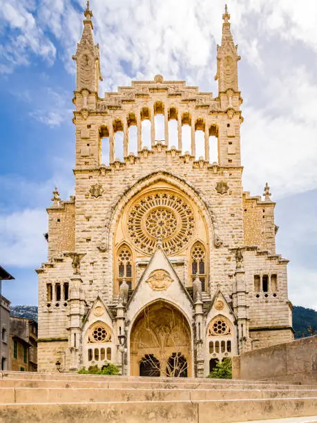 Photography of the gothic revival front facade of Sant Bartomeu Church in Soller with rose window ornaments. Staircase in the foreground. Popular tourist attraction in the old town of Sóller, Mallorca