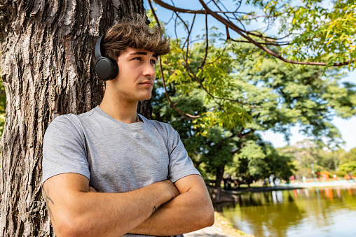 A young man listening to music, leaning on a tree with his arms folded