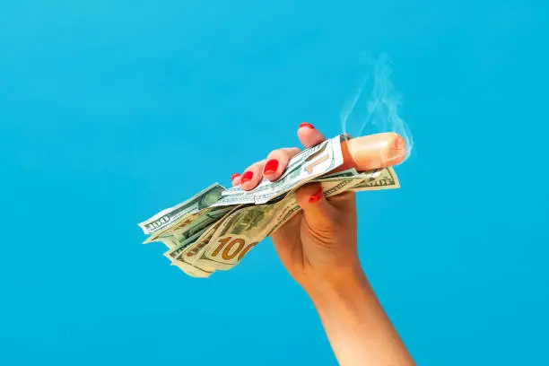 Photo of Food pop art photography. Female hand holding hot sausage wrapped in banknotes isolated on bright blue background. Vintage, retro style, surrealism, minimalism.