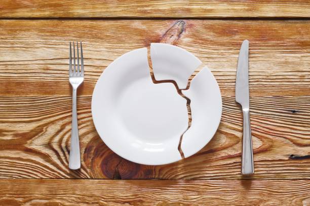 istock Top view on broken circle white plate with metal fork and knife on wooden background. The plate broke into three pieces. 1392138036