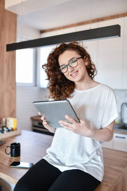 istock Cute Female Starting Work On Digital Tablet While Drinking Coffee At Home And Sitting On Kitchen Counter 1392141134