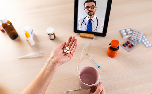 Close up of females hands holding pills and thermometer. Video call with doctor.