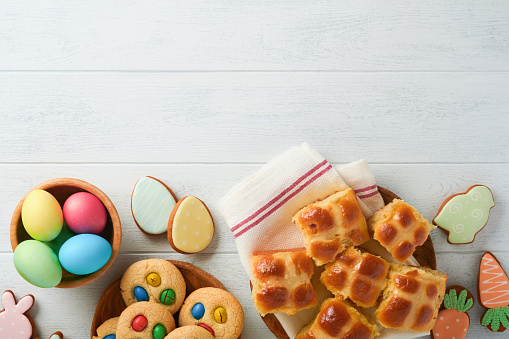 Easter hot cross buns with colored eggs, Easter gingerbread rabbits and cookies over white plank wooden old table background . Spring holiday baking concept. Easter table setting. Top view. Mock up.