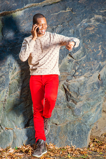 Time. Dressing in cream, patterned, collarless sweater, red jeans, boot shoes, wearing wristwatch, a young black guy is standing by rocks, smiling, making phone call, raising arm, looking at watch.