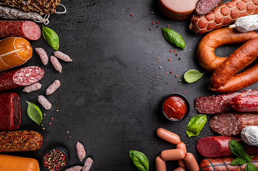 Set of different types of sausages, salami and smoked meat with basil and spices on a black stone background. Top view.