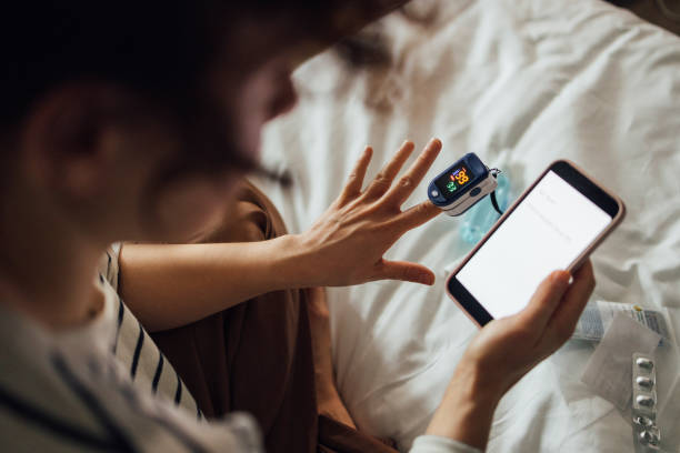 A From Above Shot Of An Unrecognizable Woman Measuring The Oxygen Saturation Level Of Her Blood After Recovering From Coronavirus While Watching Something On Her Smartphone An anonymous Caucasian female reading some health articles on her mobile phone while using oximeter to check her blood condition. medical instrument stock pictures, royalty-free photos & images