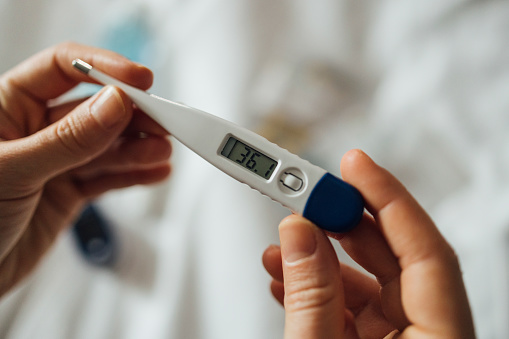 A cropped photo of an anonymous Caucasian female sitting on her bed and using digital thermometer to check her temperature.