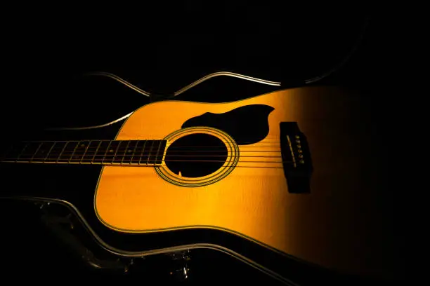 Photo of Yellow acoustic guitar lying in a hard case in the dark on a black background. Wooden stringed instrument illuminated by a beam of light. Guitar in cover close up. Rock, folklore, country music