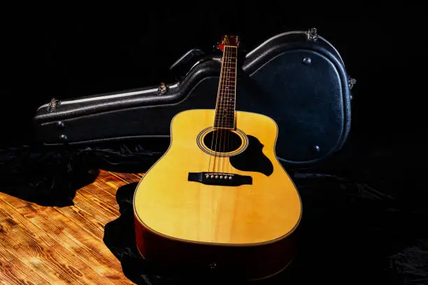 Wooden yellow acoustic guitar lying near hard case on the wooden floor. Stringed musical instrument near black open wardrobe trunk. Musical instrument with strings for performing rock, country, folklore. Close up.