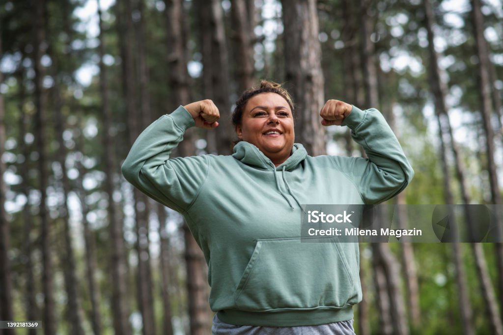 Smiling obese Hispanic Latin woman flexing arm muscles in the forest Middle-aged Hispanic woman showing arm muscles standing in forest. Obese woman exercising. Overweight woman exercising. Body positivity. Plus size woman exercising. Obesity Stock Photo