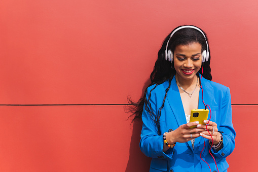 Young african woman wearing blue clothes on red background, using her mobile phone, listening to music with wireless headphones. Copy space.