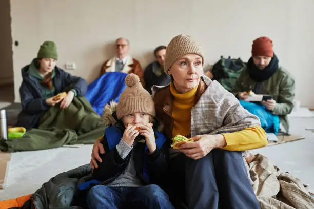 Distressed Caucasian refugee family eating sandwiches while hiding in shelter on floor covered with blankets