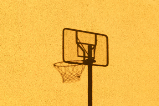 The shadow of a basketball hoop is reflected by the sunlight on a yellow house wall