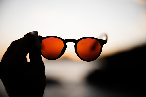 Young man holding his red - orange toned sunglasses against the setting sunset sun over Seychelles Baie Lazare Mahe Island Beach. Selective focus on sunglasses. Baie Lazare Beach, Mahe Island, Seychelles