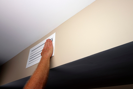 Closed white air vent of a home HVAC vent duct on a beige wall near a white color ceiling. Right hand of an adult male caucasian placed over a closed small wall rectangle air vent of a home HVAC duct