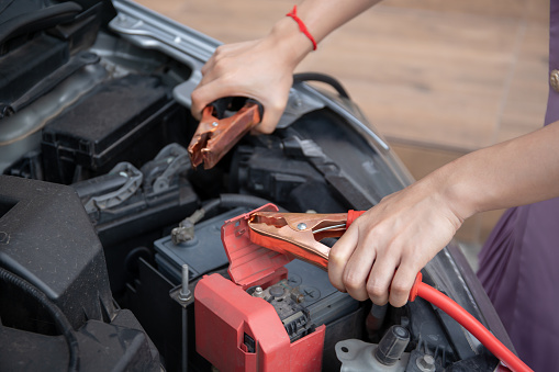 Asian young woman charging a car battery from the other car by using battery connector jump between battery. Woman showing a battery connector cable close up.